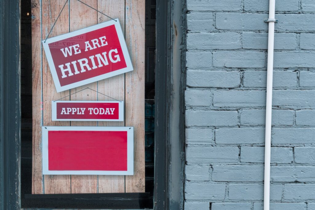 "We Are Hiring" Sign in Window of Brick Building - A red and white sign in a window of a brick building reads We Are Hiring with a smaller sign that says Apply Today.
